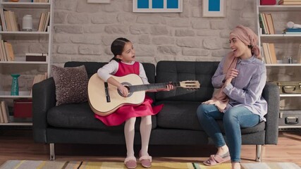 The mother with a turban is singing, her daughter is playing the guitar.Very happy smiling mother enjoy family leisure time at home with her little daughter.
