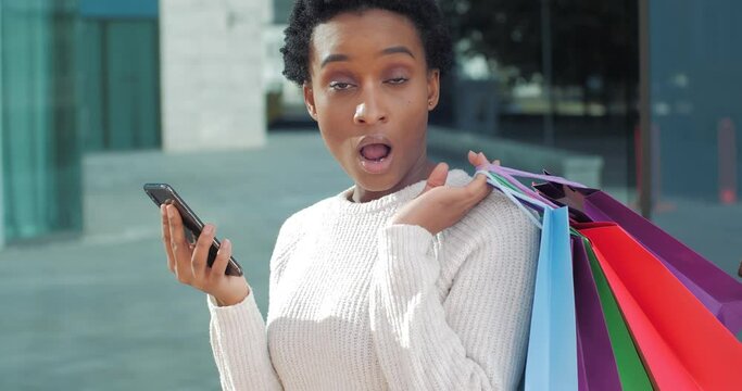 Afro american woman mixed race shopaholic girl shopper consumer looks at phone gets emeil with invoice from store feels shock surprised at high cost of new clothes and gifts holds bags of purchases