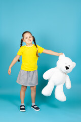 a full-length girl with pigtails in a yellow T-shirt and gray skirt holds a large white teddy bear by the ear , isolated on a blue background.