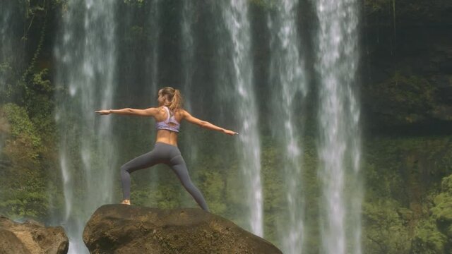 Backside view slim girl with ponytail poses in yoga exercise on large round rock against waterfall streams