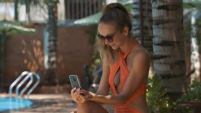 Closeup pretty long haired lady in nice orange bikini looks into mobile against swimming pool by house