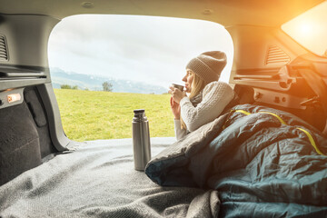 Girl resting in her car. Woman hiker, hiking backpacker traveler camper in sleeping bag, drinking hot tea and relaxing on top of mountain. Health care, authenticity, sense of balance and calmness.
