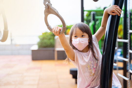 Portrait Of Young Asian Girl Wearing Mask Playing Outdoor Swinging On Monkey Bar Rings Having Fun