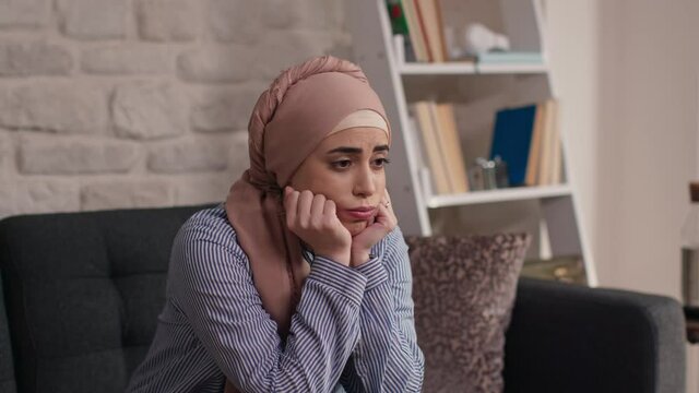 Woman in a turban sitting on the sofa at home, with her head on her hands.Portrait of thoughtful woman in headscarf sitting on sofa at home, resting her head on her hands and looking away with sad fac