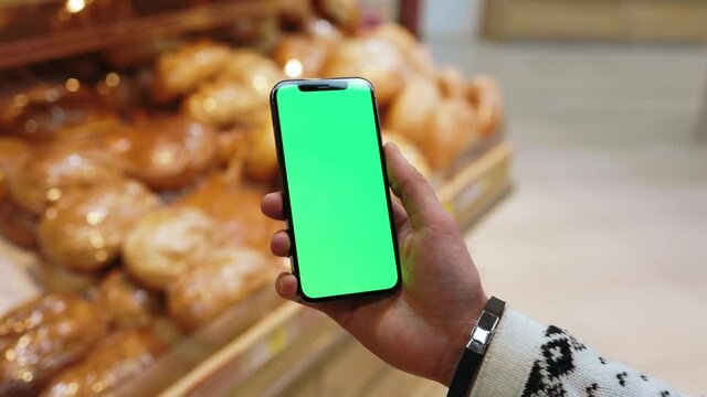 Young hands man use mobile smartphone with vertical green screen in supermarket blurred background with bread. Mockup tapping display chromakey telephone. Slow motion