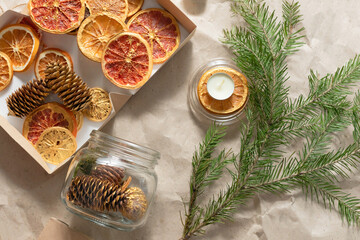 A top view of pine cones in glass can are on kraft beige paper background with dry slices of gold orange, grapefruits, green pine branch, candles. Handmade Christmas candle making concept