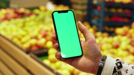 Hands man hold use mobile smartphone with vertical green screen in supermarket blurred background fruits. Mockup tapping display chromakey telephone. In store. Slow motion