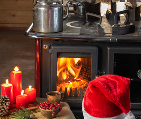 In front of a wood burning stove, a table with red candles, a cup and a red berry and a man in a red cap