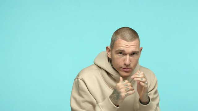 Slow motion of cool tattooed guy in beige hoodie practice jab punches, shadow boxing and looking serious at camera, standing in boxer pose over turquoise background