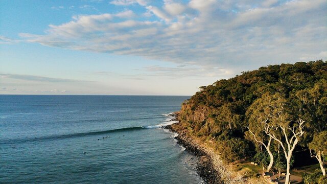 Drone photo of Noosa Heads National Park at sunset along first point. Surfers ride small waves alongside the lush forest