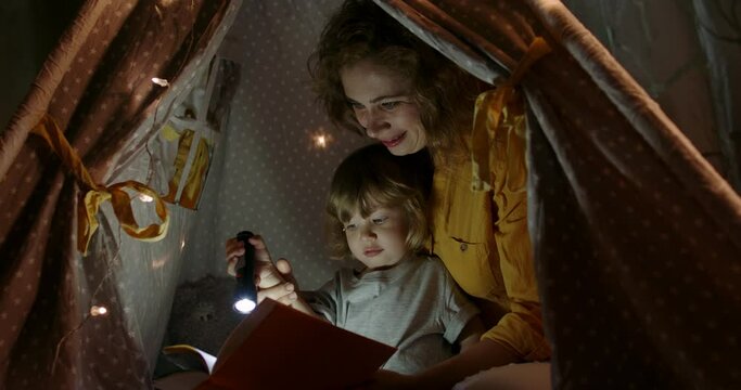 Happy caucasian mom and her cute baby boy spending time together at home, reading a book, using a flashlight in a cozy tent - happy family 4k footage