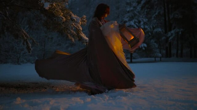 Epic fantasy shot: Young woman spinning her flying cloak holding vintage lantern in snowy forest at night. Medieval fairytale cosplay concept