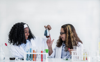 Happy african and caucasian child girl student with different skin is learning and test science chemical with colorful liquid in beaker in laboratory room at to school.