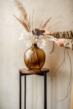 Woman Decorating Home With A Composition Of Dried Flowers And Herbs In A Glass Vase On A Beige Wall Background