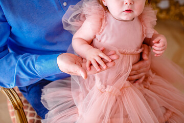Close-up of the hand of a 1 year old girl in a pink dress holding her father's fingers as she sits on his lap at home.