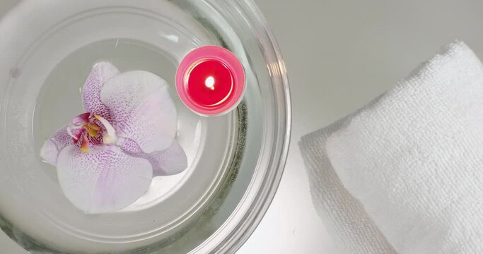 Close-up from above, a white towel is placed on a glass table, next to an archidea floating in the water and a burning candle. Interior details to create the ambience of a spa lounge zone.