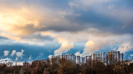 Naklejka premium Big fluffy clouds in dark blue sky over city buildings, autumn trees in foreground