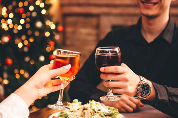 Close up of hands holding wine glasses and clinking against the backdrop of bright Christmas tree and burning fireplace, couple celebrating at Christmas table