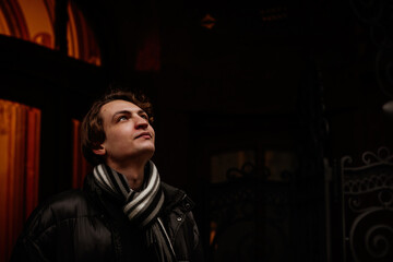 Fototapeta na wymiar Handsome stylish young man wearing winter clothes in street. Portrait of beautiful guy with modern hairstyle with jacket and scarf. Vintage European city, lights on background. Soft focus.