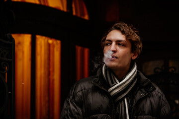 Fototapeta na wymiar Handsome stylish young hipster man smoking cigarette on the street in winter, looking away. Portrait of beautiful guy dressed casually. Vintage European city, lights on background. Soft focus.