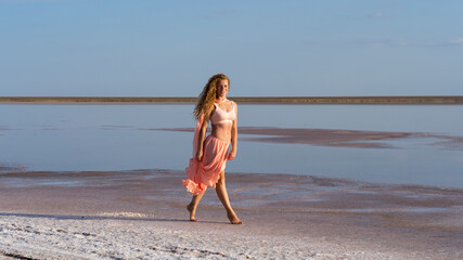 Beautiful girl in a peach dress on the white shore of a salt lake. curly model against a background of blue sky and pink salt