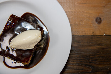 A white plate of delicious sticky toffee pudding chocolate cake with cream and chocolate sauce on a wooden kitchen work top