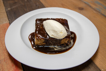 A white plate of delicious sticky toffee pudding chocolate cake with cream and chocolate sauce on a wooden kitchen work top