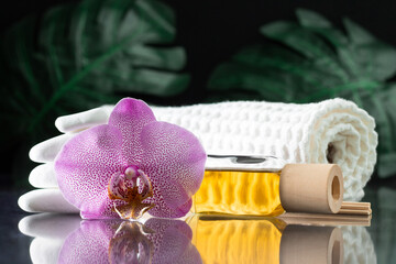 Lilac orchid flower, clear bottle of yellow oil, wooden sticks and towel with white stones and monstera on black surface