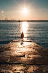 Silhouette of a person standing at the end of a jetty looking out to an industrial site filling the sky with smoke. Silhouette of a person walking on the beach during a winters sunset. 