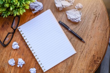 Top view of an empty notebook with eyeglasses, crumpled papers, and a pen on the wooden table 