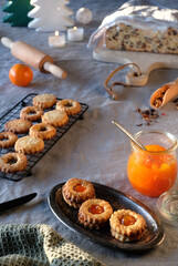 Christmas Linzer cookies filled with orange jam on cooling rack and vintage metal plate. Xmas stollen on table with natural linen tablecloth. Decorations and spices. Natural sunlight, long shadows.