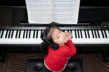 Asia toddler girl wearing headphones look up toward camera while playing piano with small smile, exploring music instrument, online learning, early childhood music education, leisure activity