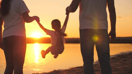 Little daughter jumping holding hands of dad and mom at sunset. Happy family life concept. A child with his parents plays together in flight. Mother and father on a walk with the kid. Spend the