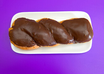 Top view flat lay of one glazed twist donut on a rectangular off white porcelain plate, on purple background.