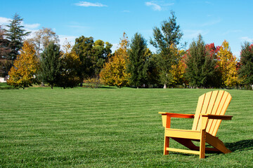 Traditional curveback sunset plastic outdoor patio adirondack chair with contoured backs and seats on green grass of manicured lawn surrouned by autumn trees. - Powered by Adobe