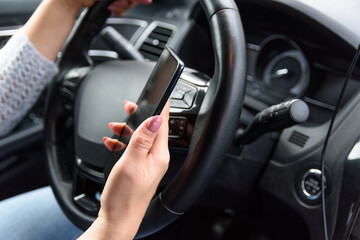 Young woman in jeans and blue sweater is driving the car with one hand, using her smartphone by another hand. Extremely unsafety and risky driving.