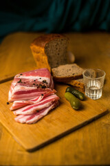 Raw bacon on a wooden plate wish shot glass, pickle and black bred. Homemade bacon on wooden board