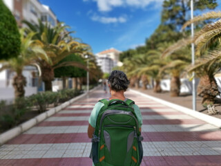 Back view of a senior woman grey hair walking in the city street with backpack on shoulders. Palm trees and blue sky on background