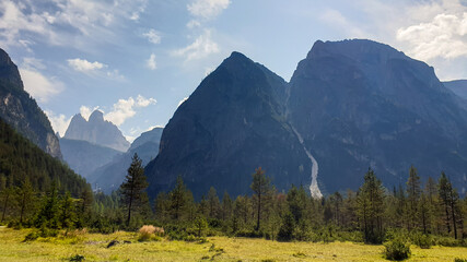 Panoramic view on the high Italian Dolomites peaks. In the back there are Drei Zinnen visible. Many high mountains around. A lush green meadow in front with a few pine trees in between. Sunny day.