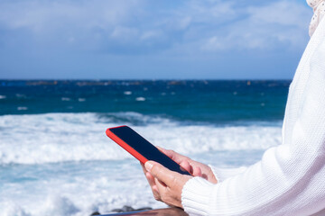 A senior woman white dressed enjoying beach vacation on a windy day reading a message in her smartphone.  Relaxed smiling active retiree