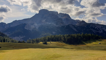 A panoramic view on the high Italian Dolomites from the top of Strudelkopf. There is a wide gravelled path leading to the top. Sunny day. A few clouds above the high peaks. Lush green plateau around
