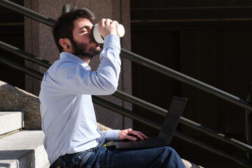 Young business man drinking coffee while is using his laptop sitting on stairs. Businessman in a urban street of a city.
