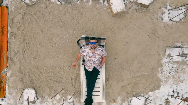 Aerial view on top worker rested in the winter on the construction site lying on a deck chair among the foundations of the house. A man drinks juice and lies on an imaginary beach among the