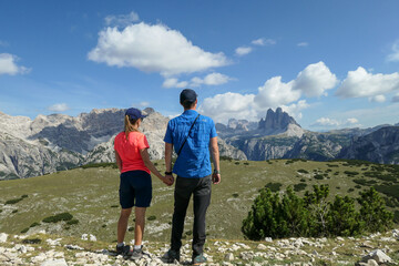 A couple holding hands and enjoying the distant view on Drei Zinnen from Strudelkopf in Italian Dolomites. A lush green meadow in front. The man enjoys the view and the trek. High Alpine landscape