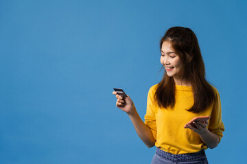 Young Asia lady using phone and credit bank card with positive expression, smiles broadly, dressed in casual clothing and stand isolated on blue background. Happy adorable glad woman rejoices success.