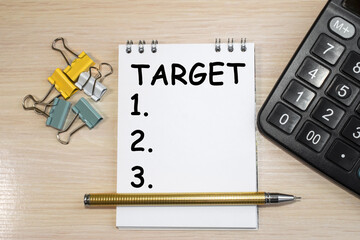 The target is written on a notepad with a white sheet and a pen that lies on a wooden surface among a calculator and colored paper clips.