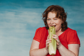 Cute smiling chubby red-haired girl bites celery and smiles against a blue background.