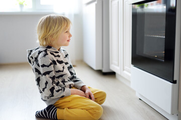 Glad child sitting on the floor near kitchen stove and waiting for a pie or other baked goods to be prepared