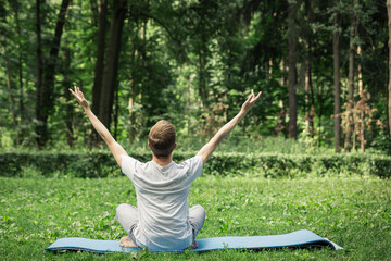 Young attractive man in sport clothes is meditating in the lotus position in the park against the background of grass and trees. Hands up to the sky