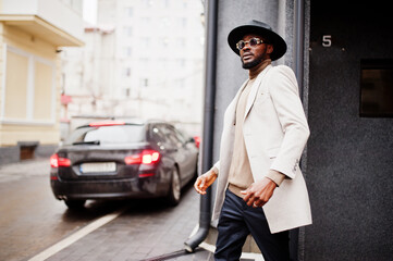 Stylish african american man wear beige jacket and  black hat with sunglasses pose near car.